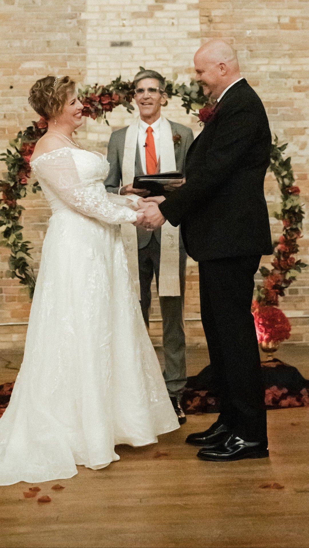 Randy Johncox officiating a wedding ceremony as the bride and groom hold hands and smile at each other before a floral arch in a brick venue