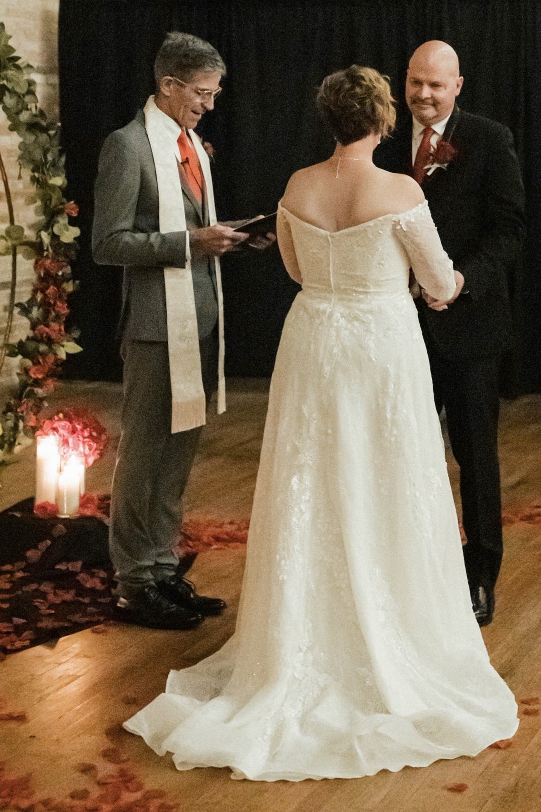 Randy Johncox reading during a wedding ceremony, the bride in a lace gown facing the smiling groom beside glowing candles