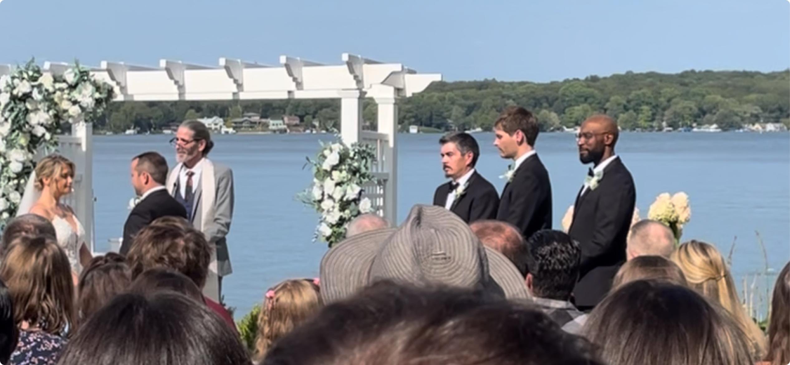 Randy Johncox officiating a lakeside wedding ceremony, the couple beneath a white flower-draped pergola with the lake behind them